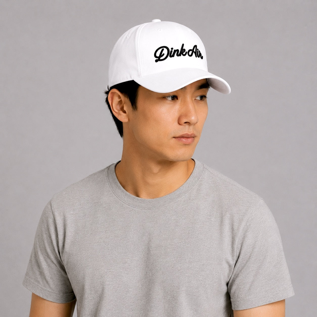 Male model wearing the white Dink Air Script Hat featuring the embroidered Dink Air logo, styled with a light gray performance tee against a neutral studio background that showcases the hat’s clean, modern design.