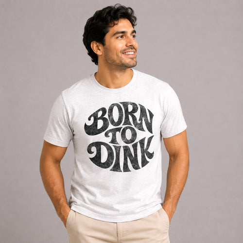 Male model wearing the Born to Dink Premium Tri-Blend Tee in heather white with a distressed black "Born to Dink" graphic, paired with khaki pants in a clean studio setting, showcasing the ultra-soft premium tri-blend fabric and relaxed athletic fit.