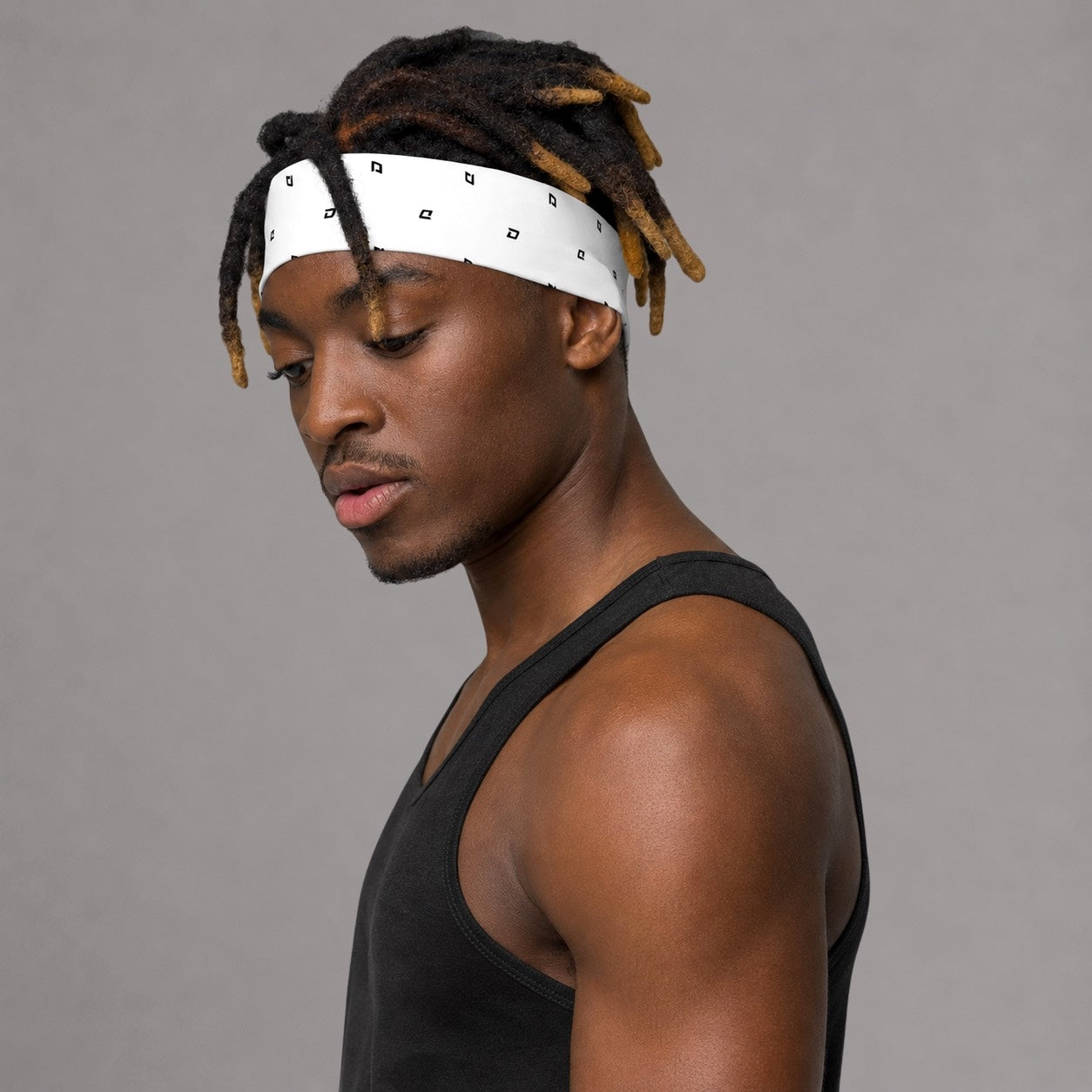 Black male model wearing the D Mark Performance Headband in white with an all-over D Mark pattern, paired with a black athletic tank in a clean studio setting, showcasing the lightweight, moisture-wicking performance fabric and comfortable athletic fit.