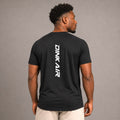 Male model wearing the Dink Air Vertical Performance Tee in black, shown from the back with the vertical white Dink Air logo in a neutral studio setting.