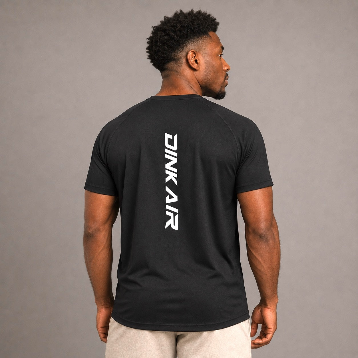 Male model wearing the Dink Air Vertical Performance Tee in black, shown from the back with the vertical white Dink Air logo in a neutral studio setting.