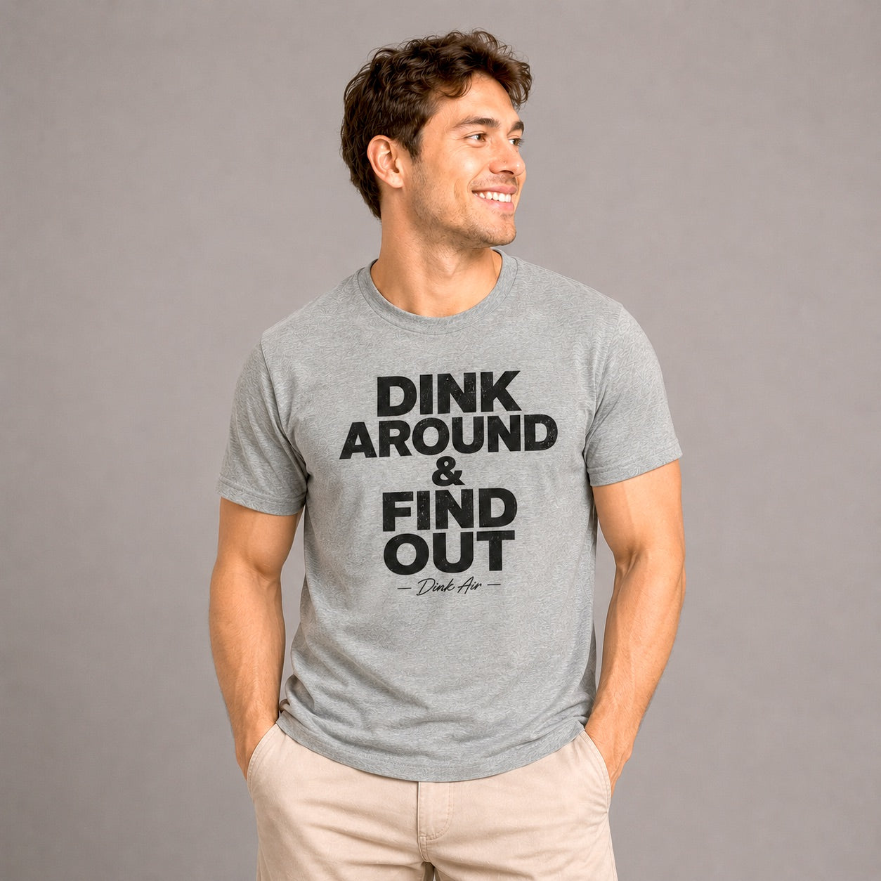 Male model wearing the Dink Around & Find Out Premium Tri-Blend Tee in heather gray with a bold black statement graphic, paired with light khaki pants in a clean studio setting, showcasing the ultra-soft premium tri-blend fabric and relaxed athletic fit.