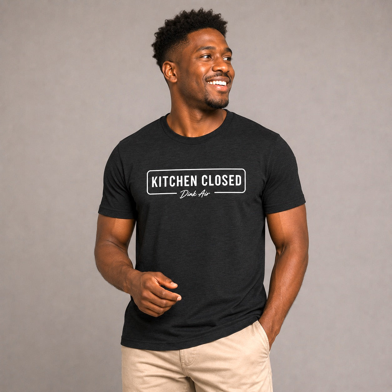 Male model wearing the Kitchen Closed Premium Tri-Blend Tee in black with a clean white "Kitchen Closed" graphic and Dink Air script, paired with light khaki pants in a neutral studio setting that highlights the ultra-soft premium tri-blend fabric and relaxed athletic fit.