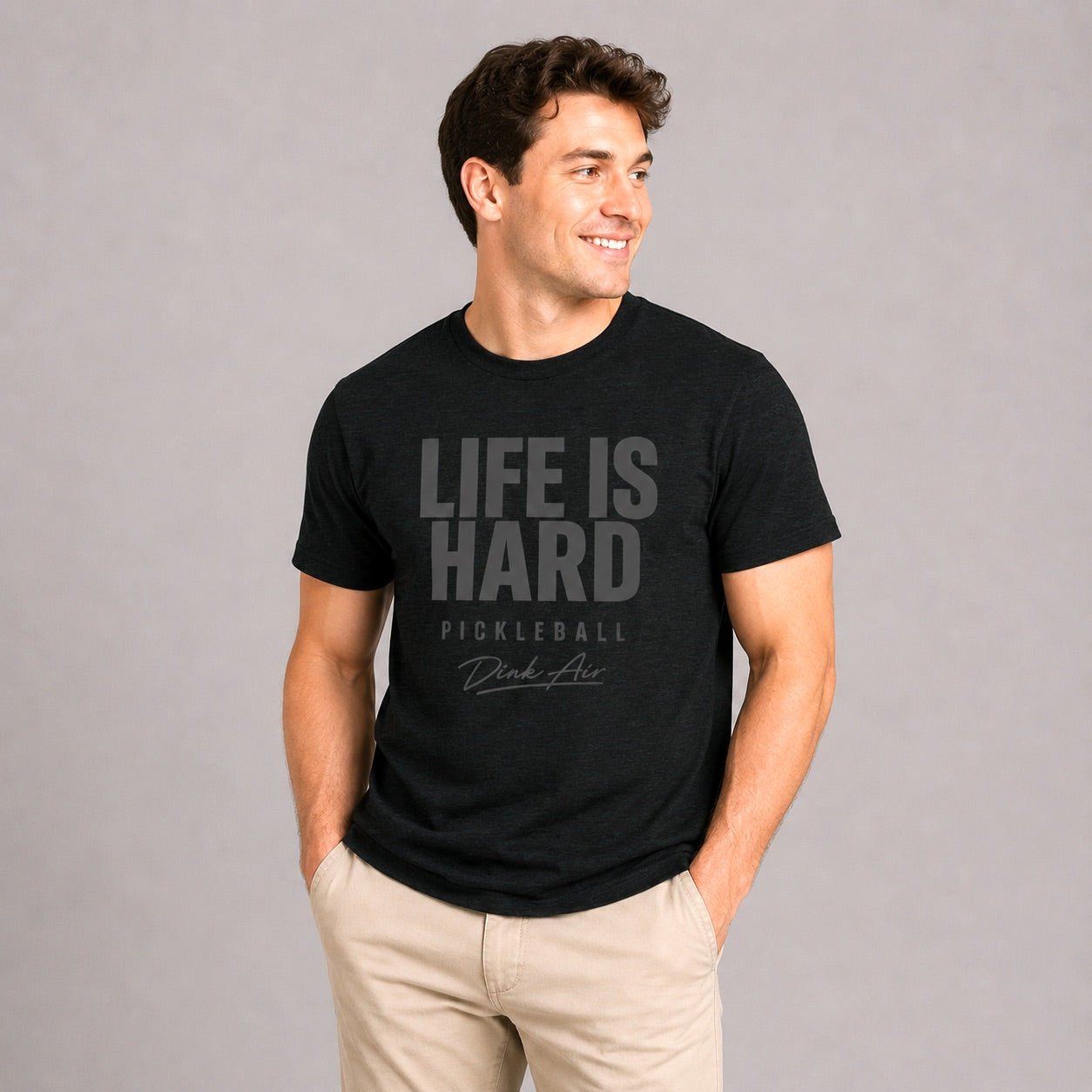 Male model wearing the Life Is Hard Tee in vintage black with a distressed "Life Is Hard Pickleball" graphic, paired with light khaki pants in a clean studio setting, showcasing the ultra-soft premium tri-blend fabric and relaxed athletic fit.