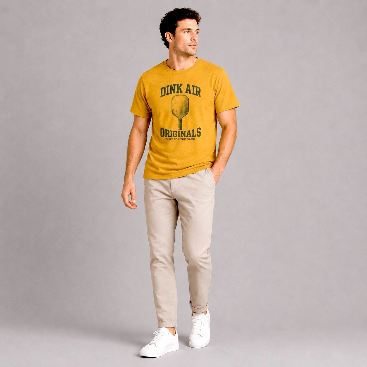 Male model wearing the mustard Originals Heritage Tee featuring a vintage-inspired pickleball graphic, styled with light-colored pants in a casual lifestyle setting.