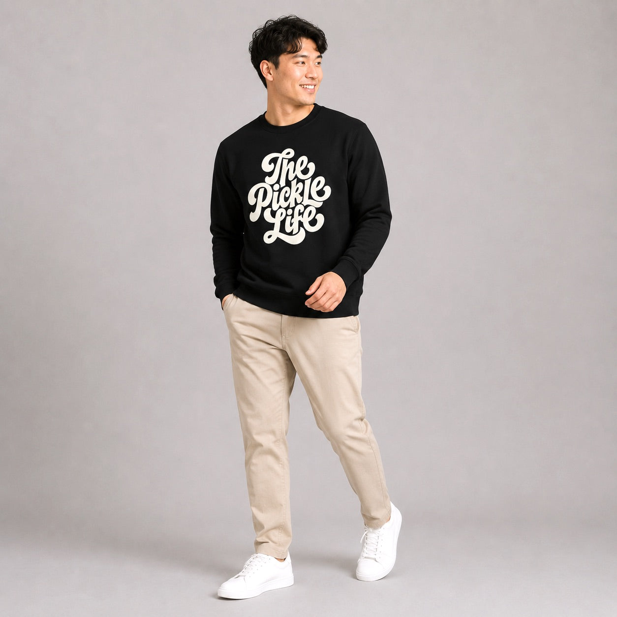 Male model wearing the Pickle Life Premium Crewneck sweatshirt featuring a bold retro-inspired "The Pickle Life" graphic, paired with light khaki pants and white sneakers in a clean studio setting that highlights the sweatshirt's premium comfort, relaxed fit, and timeless pickleball lifestyle appeal.
