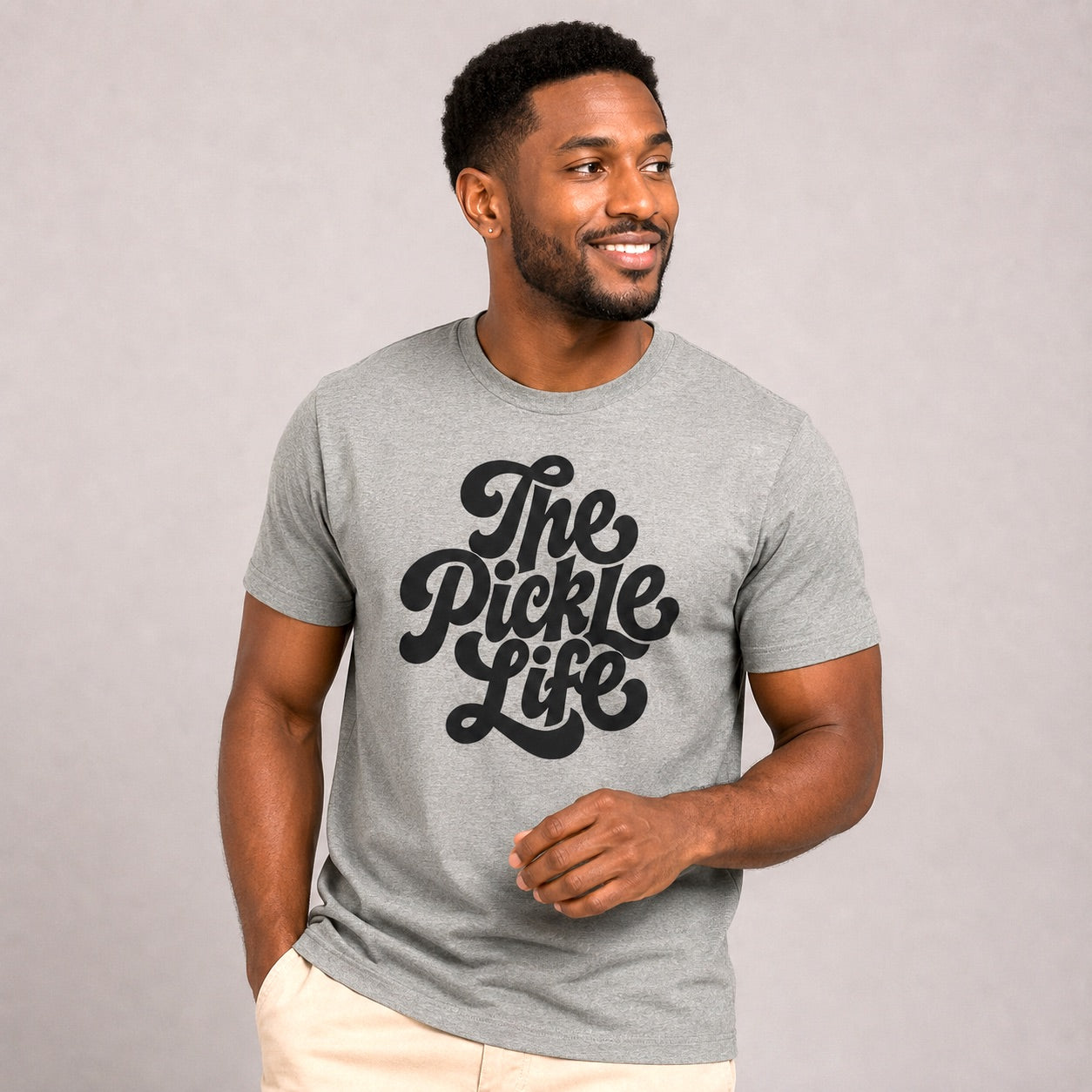 Male model wearing the Pickle Life Premium Tee in heather gray with a bold vintage-inspired black "The Pickle Life" graphic, paired with light khaki pants in a clean studio setting that highlights the ultra-soft premium tri-blend fabric, relaxed athletic fit, and timeless pickleball lifestyle style.
