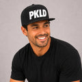 Smiling man wearing the PKLD Pickled Snapback Hat featuring bold white PKLD embroidery on a black flat-brim snapback. Premium pickleball lifestyle cap styled with a black t-shirt in a clean studio setting, designed for comfort on and off the court.